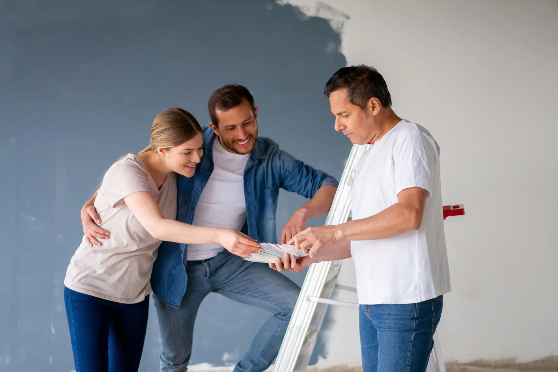 Painter reviewing a colour fan deck with a couple beside a half-painted blue-grey wall
