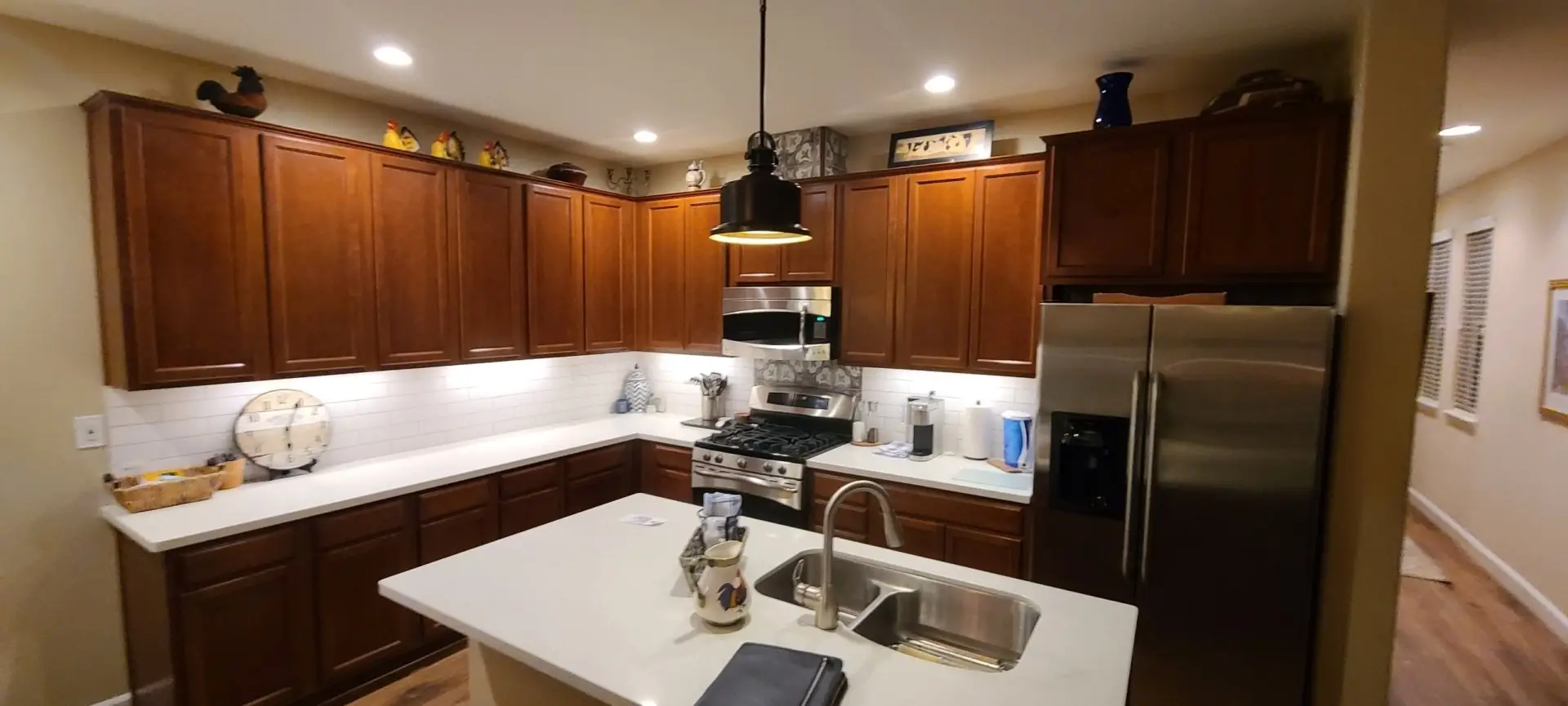 Wide view of an L-shaped kitchen with wood-stained cabinets, stainless appliances and a pendant-lit island