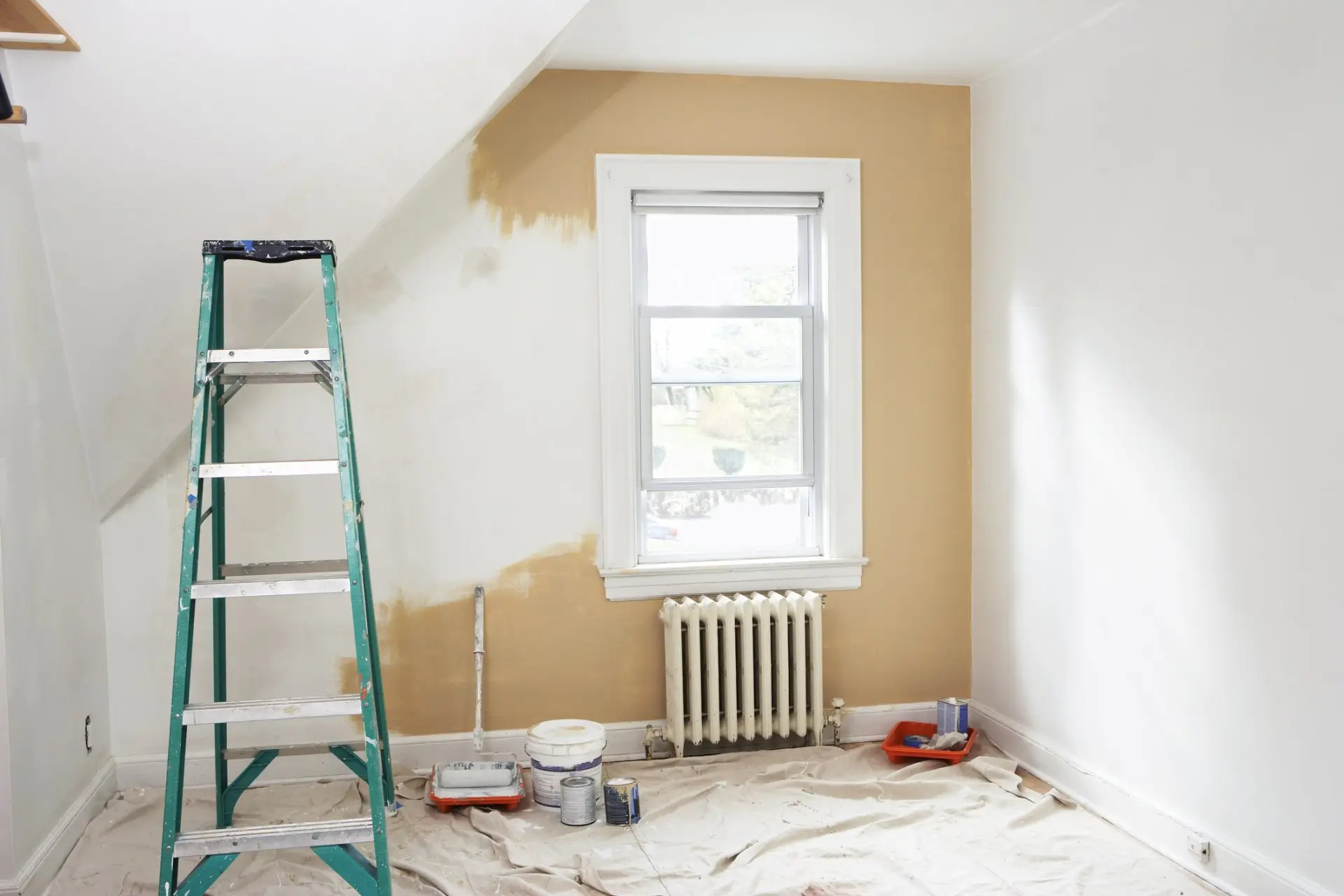 Bedroom mid-repaint with a tan accent wall, stepladder, drop cloths and paint supplies